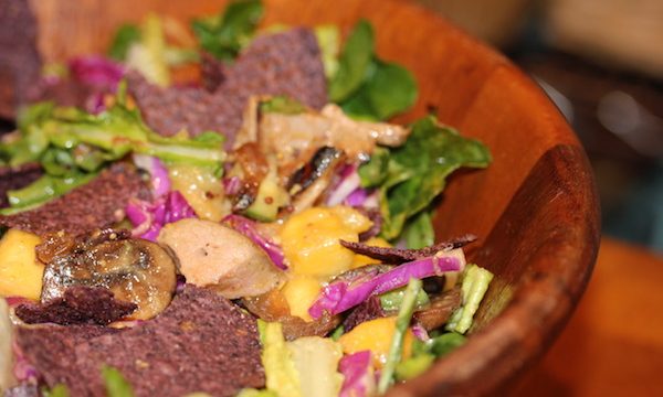 Large wooden salad bowl filled with salad greens, red cabbage ribbons, cubes of pork and blue corn tortilla chips