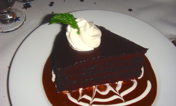White plate with very dark chocolate cake slice sitting in a pool of chocolate sauce and white creme anglaise sauce in a spider web pattern and a swirl of whipped cream on top with 1 mint leaf