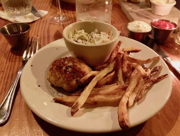 A white plate with a crab cake, fries and a small bowl of slaw