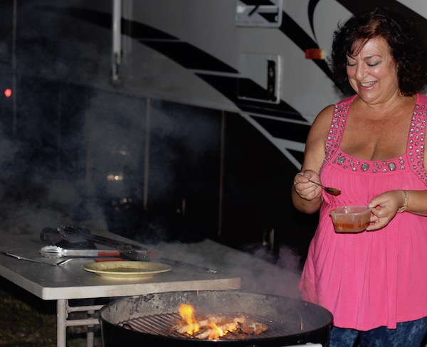 A dark haired woman with a hot pink top brushing skewered shrimp with butter on a hot grill.