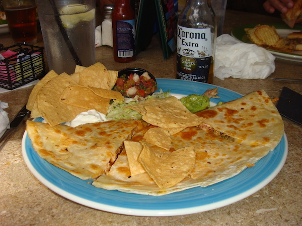 A blue rimmed round plate topped with a quesadilla, tortilla chips and guacamole with a bottle of Corona Extra in the background