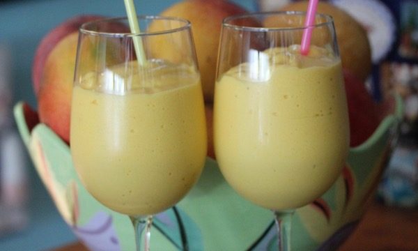 Two wine glasses with pale yellow smoothies and straws in front of a painted ceramic bowl filled with ripe mangoes.
