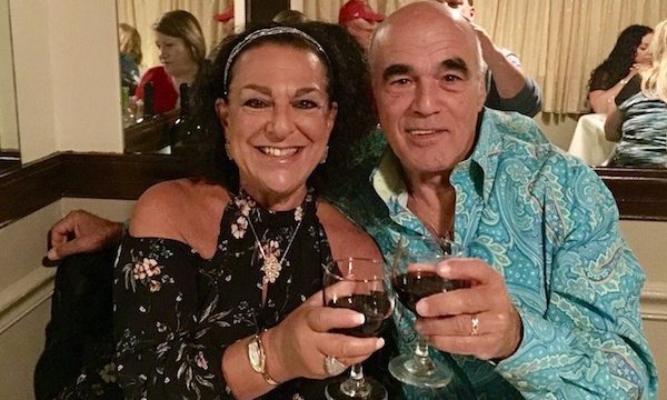 Man in turquoise shirt and woman in black blouse toasting with glasses of red wine