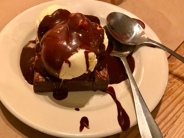 A white plate with a brownie square topped with a ball of vanilla ice cream and covered with Hot Fudge and two spoons