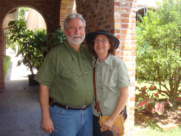A bearded grey haired man and a woman in a brimmed hat in front of a brick column.