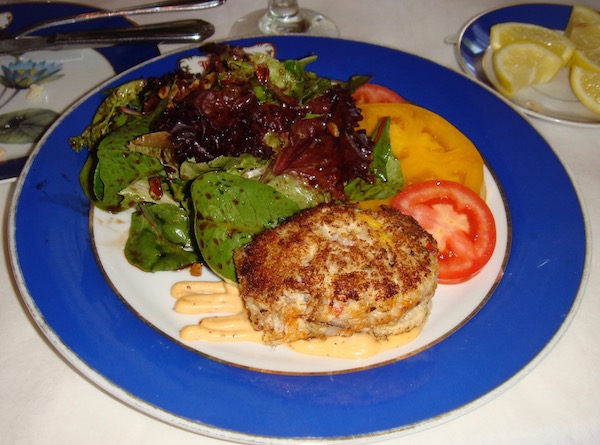 A dinner plate with a wide cobalt blue rim topped with a crab cake, salad greens and sliced tomatoes