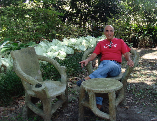 A bald headed man in a red t-shirt and jeans relaxing in a garden on a chair that looks like it is made of trees.