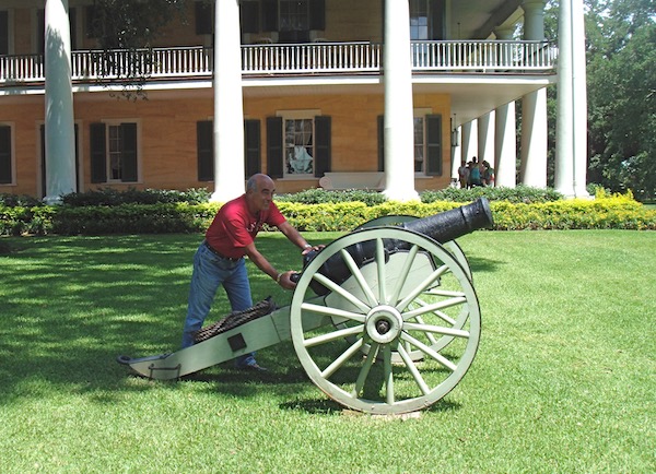 A man in a red t-shirt and jeans pretending to shoot a canon on a lawn.