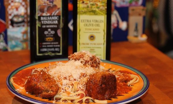 A yellow and blue pasta dish filled with spaghetti, 3 meatballs and grated parmesan sitting on a butcher block with bottles of olive oil and balsamic vinegar behind it.