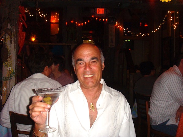 Smiling man in white shirt sitting in restaurant holding up a martini with olives