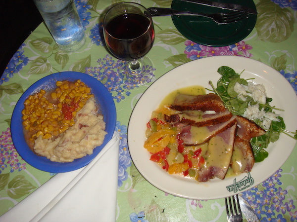Floral tablecloth set with a small blue bowl of mashed potatoes and vegetables along side a white plate with tuna slices fanned on plate and a citrus relish with a glass of red wine in between