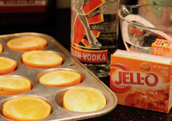 A muffin tin filled with hollowed orange rind halves, a bottle of vodka, a measuring cup with water and two boxes of orange jello mix.