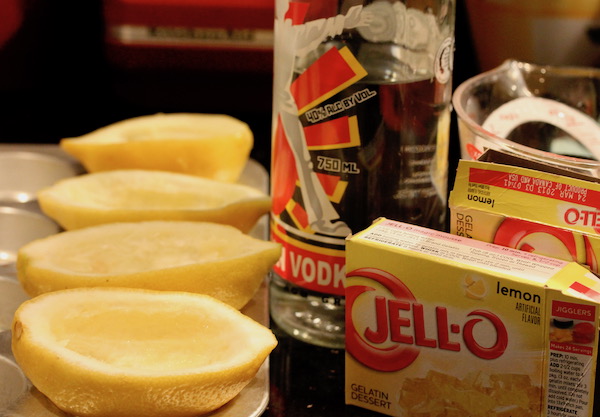 A muffin tin filled with hollowed lemon rind halves, a bottle of vodka, a measuring cup with water and two boxes of lemon jello mix.