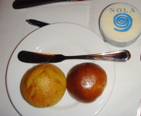 A white bread and butter plate topped with two rolls and a butter spreader with a small bowl of butter in upper right