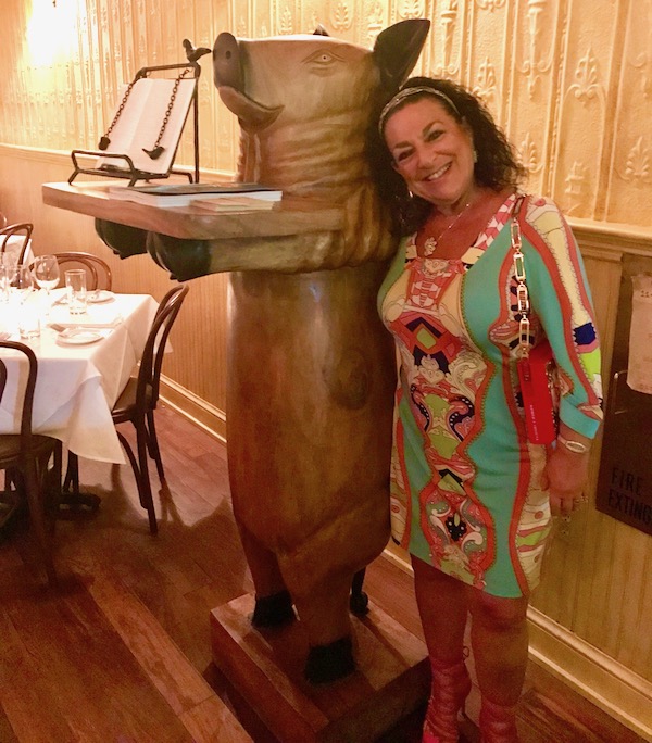 A dark haired woman in a print dress Posing with La Petite Grocery’s Mascot Pig