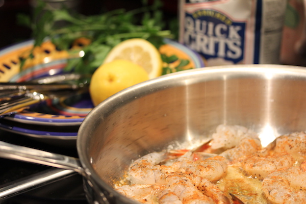 A steel pan with shrimp cooking in oil. Behind pan is a lemon cut in half, some parsley and a bag of Quick Grits