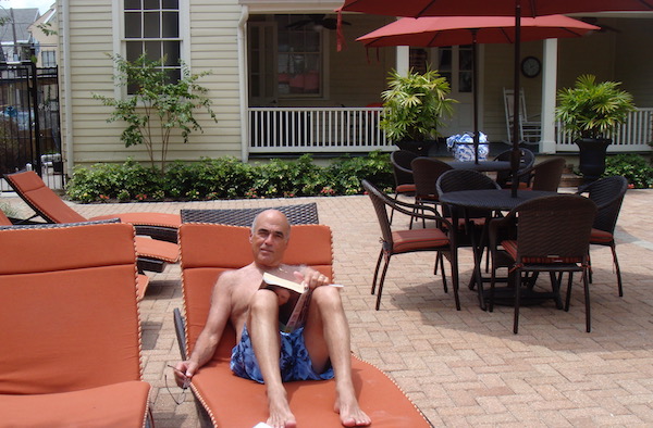 A bald man in bathing trunks relaxing on a chaise lounge with a book on a patio.
