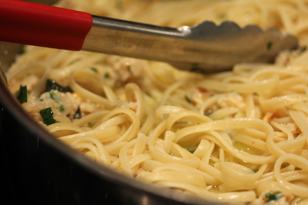 A steel pan withLinguine with White Clam Sauce cooking and being stirred by a pair of red handled tongs