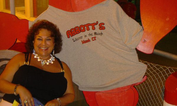 Dark haired woman in a black top with white pants leaning against a giant red lobster with white t-shirt on