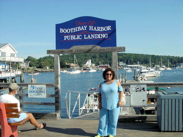 A dark haired woman in light blue top standing on dock in front of Boothbay Harbor sign