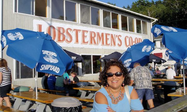 A dark haired woman in light blue top leaning agains a wood rail at an outdoor café