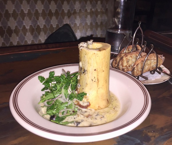 A white plate with a large hollow bone standing on it and sauce with marrow and snails