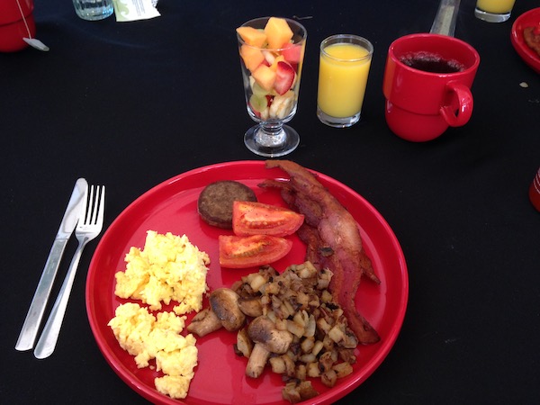 A large red plate filled with scrambled eggs, sausage patty, bacon, 2 tomato wedges and home fries on a black tablecloth along with a glass of fresh fruit, a glass of orange juice and a red mug with black coffee.