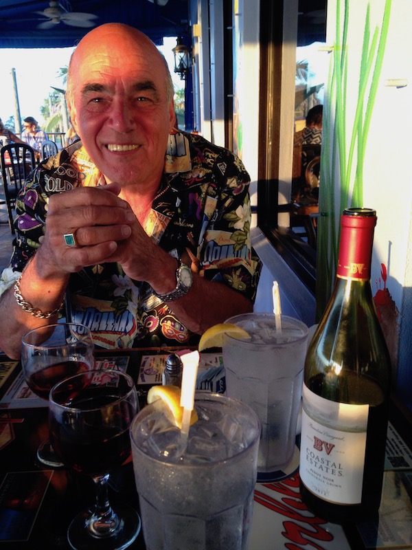 A smiling man in a tropical shirt sitting at a restaurant table with a bottle of wine