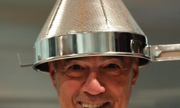 A man smiling and holding a cone shaped strainer on his head