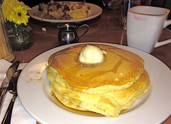 A white plate topped with a tall stack of pancakes, a pat of butter and syrup.