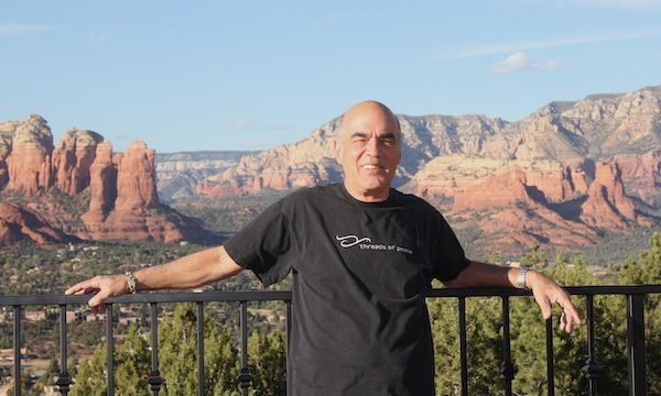 A bald man in a black t-shirt leaning against a rail with red rock formations in the background.