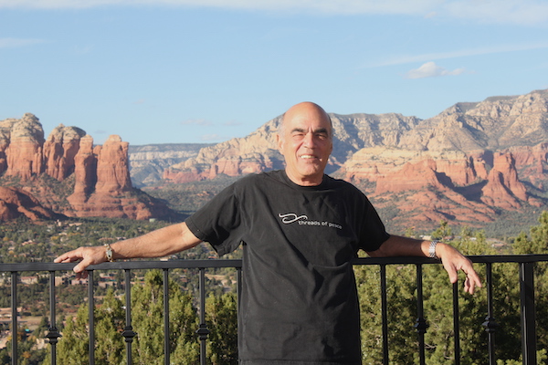 A bald man in a black t-shirt leaning against a rail with red rock formations in the background.