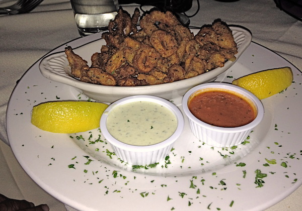 An oval white bowl filled with fried calamari sitting on a white platter with two ramekins holding a white sauce and a red sauce.