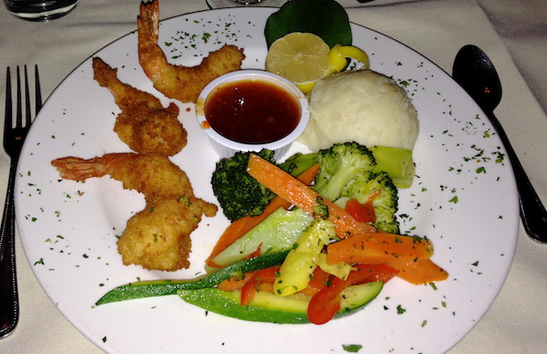 A white dinner plate topped with three fried shrimp, vegetables and mashed potatoes with a lemon half sprinkles with chopped parsley