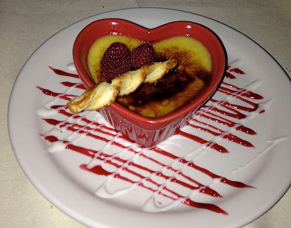 A heart shaped red dish with custard sitting on a white plate with a cookie straw and red sauce drizzled on the plate and custard.