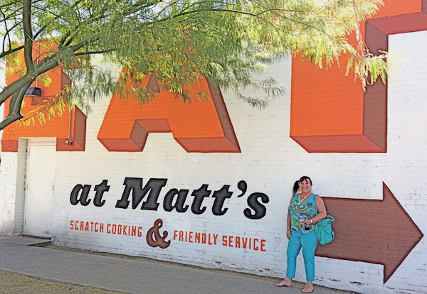 A woman in a turquoise outfit in front of a wall painted with Eat at Matt's and an arrow pointing the way.