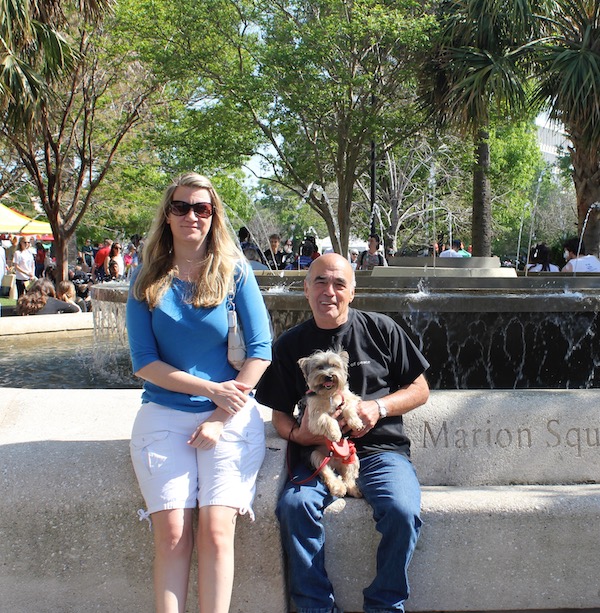 A young woman with long blond hair and an older man holding a Yorkie sitting on a low wall at a park