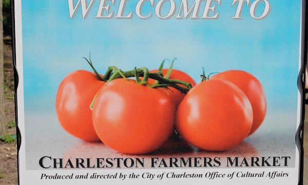 A sign Welcome to Charleston Farmers' Market with a bunch of ripe red tomatoes on the vine against an aqua background.