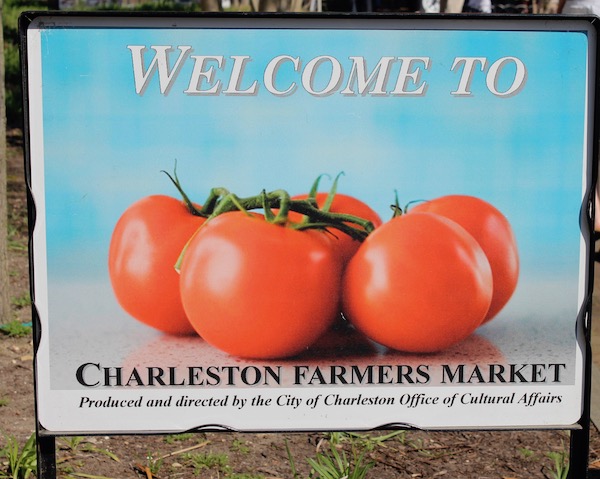 A sign Welcome to Charleston Farmers' Market with a bunch of ripe red tomatoes on the vine against an aqua background.