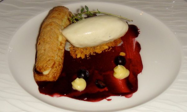 A white plate topped with a long profiterole, a vanilla ice cream quenelle, blackberry sauce and fresh fruit.