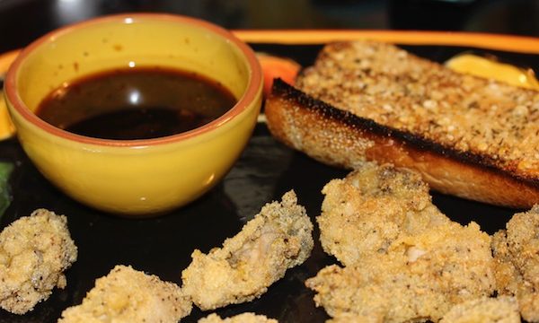 A black platter with fried oysters, a small yellow bowl with a brown sauce and a slice of garlic bread top right
