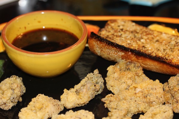 A black platter with fried oysters, a small yellow bowl with a brown sauce and a slice of garlic bread top right