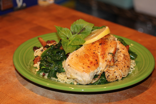 Olive green dinner plate with pan seared chicken breast leaning a top angel hair pasta which is topped with wilted spinach and garnished with basil leaves
