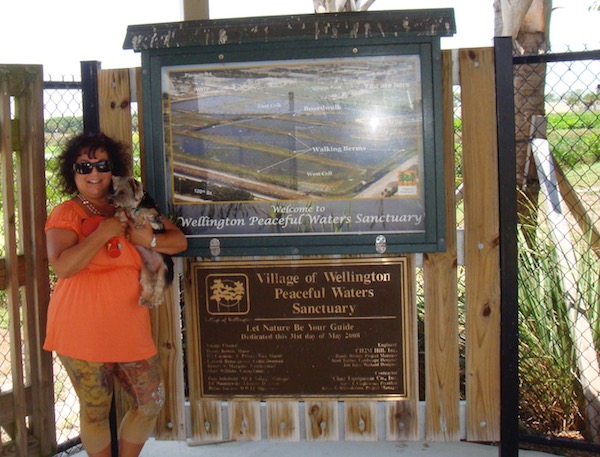 A woman in a coral top holding a Yorkie to the left of a sign for Peaceful Waters Sanctuary