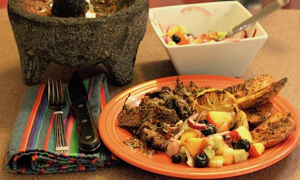 A molcajete with red salsa, an orange plate with Grilled Skirt Steak with Red Chimichurri and Peach Blueberry Tomatillo Relish and a square white bowl with the fruit relish and a spoon