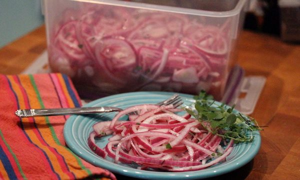An aqua plate filled with pickled red onions and a fork with a striped napkin to the left and a plastic container filled with pickled red onions behind.