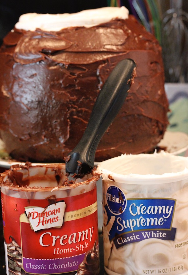 A container of Duncan Hines Chocolate Frosting and one of Pillsbury Classic White frosting with chocolate frosted cake behind.