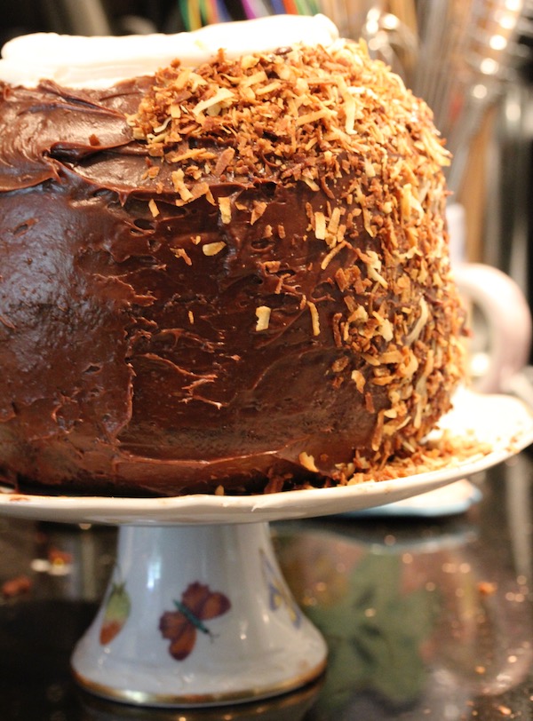 A pineapple shaped cake frosted in chocolate and partially covered with toasted coconut on a white cake stand