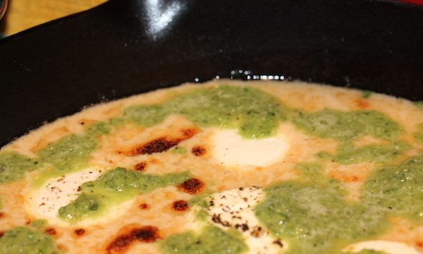 A cast iron pan filled with melted cheese dip with brown spots and green sauce