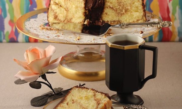 A partial yellow cake with chocolate frosting on a gold rimmed glass cake stand with a slice of cake in front and a black mug of coffee
